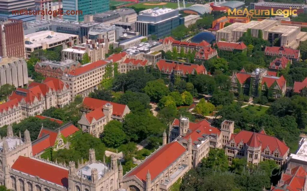 An aerial view of a university campus featuring Gothic-style stone buildings with red roofs surrounded by lush green trees.