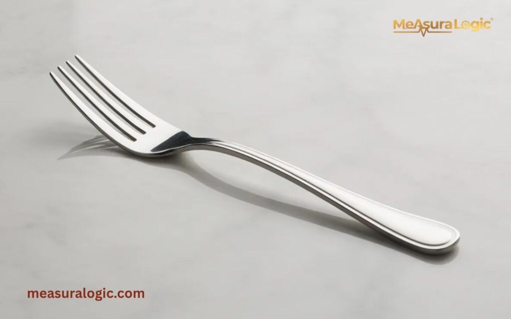 A salad fork lies horizontally on a greyish-white surface. The fork features four long tines and a simple, rounded handle with a classic ridged border.