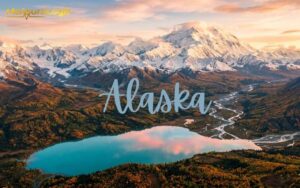 An aerial view of Alaskan wilderness at golden hour, featuring a lake. Winding glacial rivers cut through vibrant autumn forests toward the towering, snow-capped peak of Denali.
