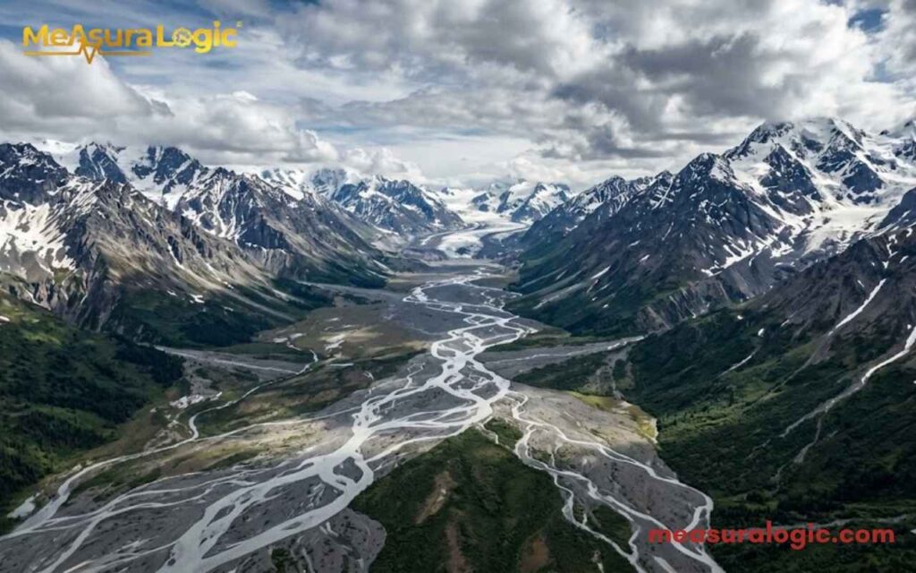 An epic aerial panorama of a vast glacial valley flanked by snow-capped mountains.
A complex braided river snakes through the valley floor under a dynamic cloudy sky.