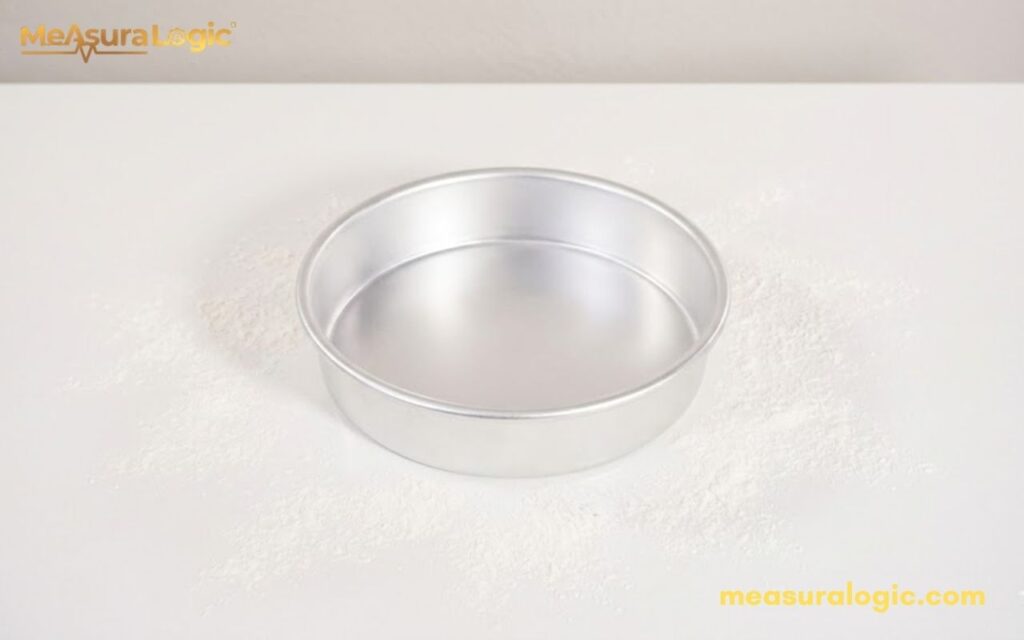 A shiny silver 9 inches round cake pan centered on a clean white surface. A light dusting of flour surrounds the pan.