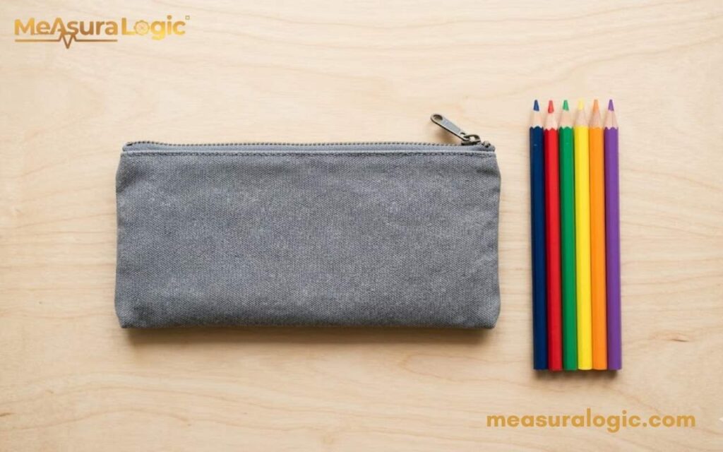 A view of a 9 inches gray zippered pencil case resting on a light wooden desk. Six brightly colored pencils are neatly aligned right next to it.