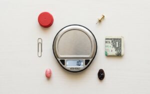A digital scale reading 1.0g sits centered on a white surface. A paperclip, bottle cap, thumbtack, folded dollar, raisin, and jelly bean surround it.